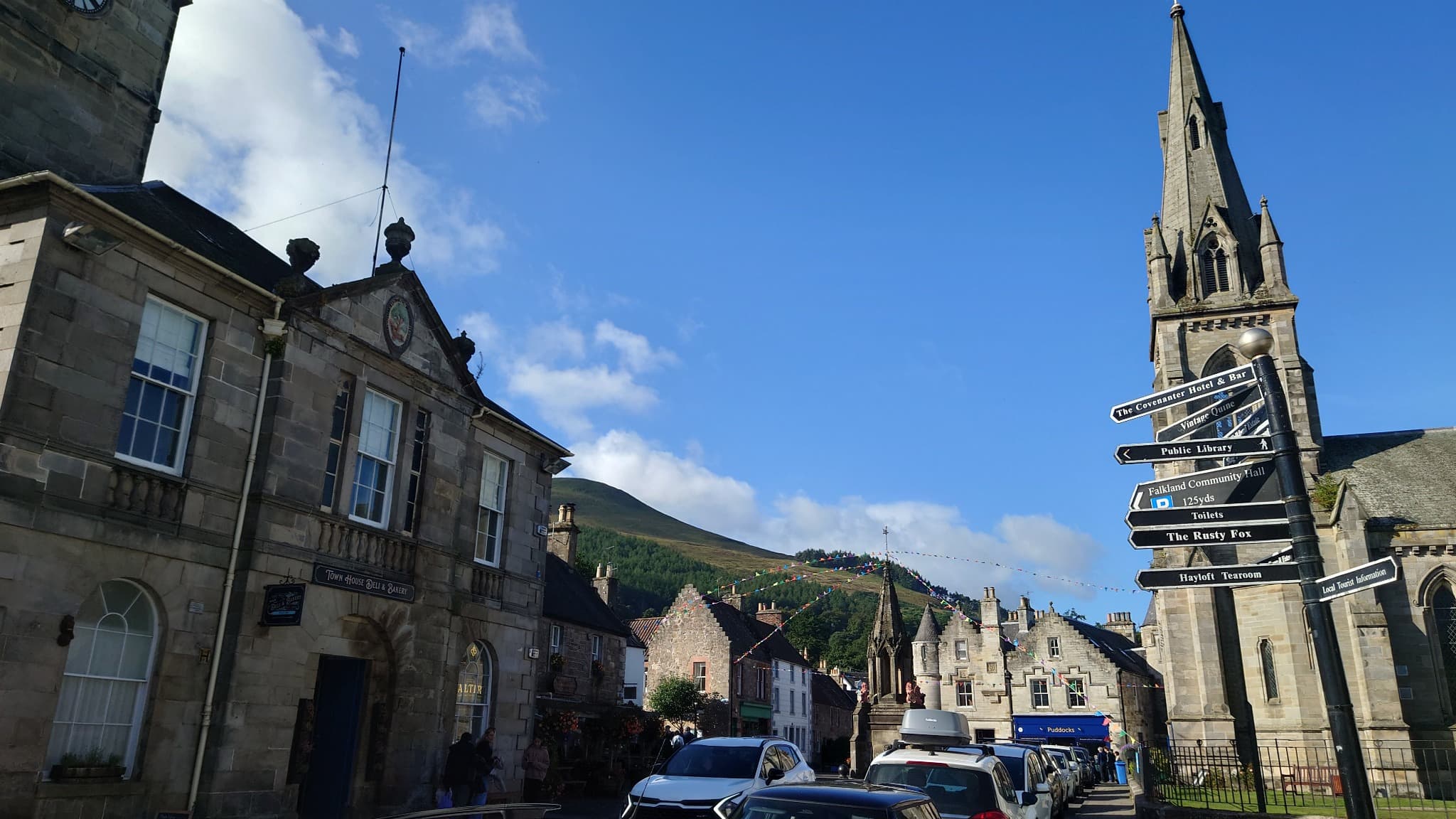 Falkland village with historic stone buildings, church spire and direction signs beneath a blue sky.